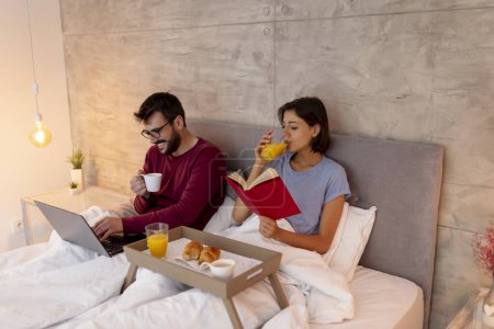 Photo for Beautiful young couple in love wearing pajamas, having fun reading and surfing the net using laptop computer while having breakfast in bed - Royalty Free Image