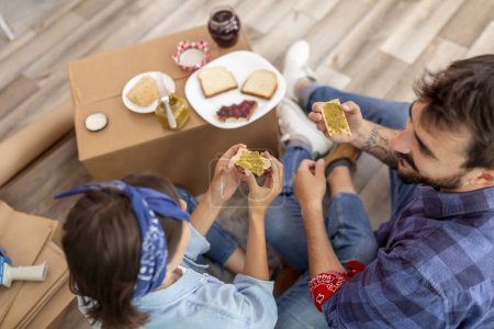 Foto de Vista superior de la joven pareja enamorada sentada en el suelo entre cajas de cartón, divirtiéndose mientras desayunan en su nuevo apartamento - Imagen libre de derechos