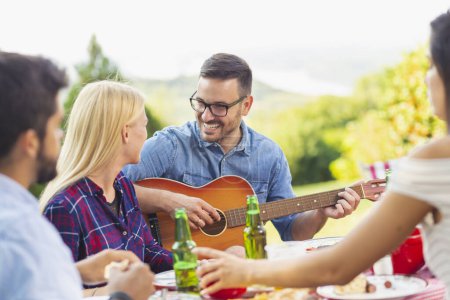 Téléchargez les photos : Groupe d'amis faire une fête barbecue à l'extérieur, boire de la bière, griller de la viande, jouer de la guitare, chanter et s'amuser - en image libre de droit