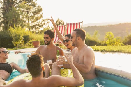 Téléchargez les photos : Groupe d'amis à une fête d'été au bord de la piscine, s'amuser dans la piscine, boire des cocktails et de la bière et porter un toast - en image libre de droit