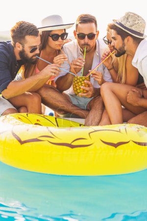 Téléchargez les photos : Groupe d'amis à une fête d'été au bord de la piscine, assis au bord d'une piscine, boire un cocktail d'ananas et s'amuser - en image libre de droit