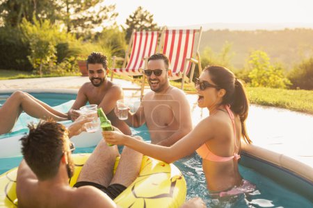 Téléchargez les photos : Groupe d'amis à une fête d'été au bord de la piscine, s'amuser dans la piscine, faire un toast et boire des cocktails et de la bière - en image libre de droit