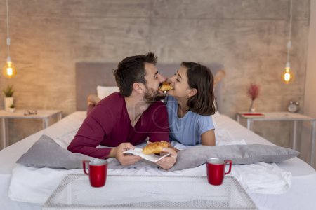 Téléchargez les photos : Couple amoureux en pyjama, couché au lit, petit déjeuner et café au lit après s'être réveillé le matin. Couple partageant un croissant - en image libre de droit