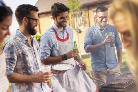Photo for Group of friends making barbecue in the backyard, drinking beer and having fun on a sunny summer day. Focus on the man in the middle - Royalty Free Image