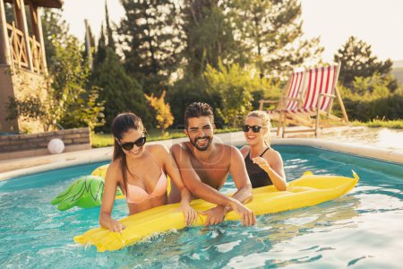 Foto de Grupo de amigos en una fiesta de verano junto a la piscina, divirtiéndose en la piscina, salpicando agua y peleando por un colchón flotante - Imagen libre de derechos