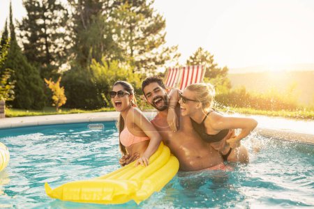 Téléchargez les photos : Groupe d'amis à une fête d'été au bord de la piscine, s'amuser dans la piscine, éclabousser de l'eau et se battre sur un matelas flottant - en image libre de droit