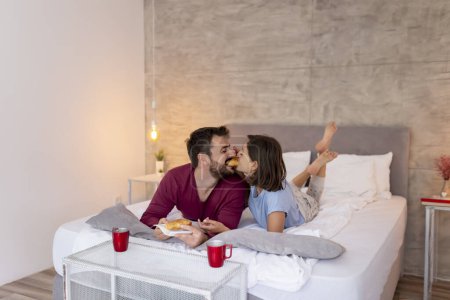 Photo for Couple in love wearing pajamas, lying in bed, having breakfast and coffee in bed after waking up in the morning. Couple sharing a croissant, biting it from both ends - Royalty Free Image