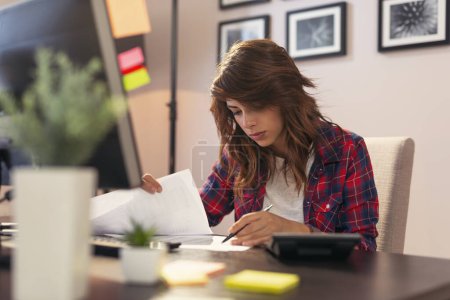 Foto de Mujer joven revisando documentos, trabajando hasta tarde en una oficina en casa - Imagen libre de derechos