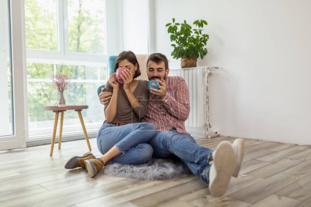 Téléchargez les photos : Beau jeune couple amoureux assis sur le sol de leur nouvelle maison, embrasser, rire et boire du café - en image libre de droit