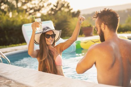 Photo for Couple in love at a poolside summer party, standing in water next to the swimming pool edge, drinking beer and having fun - Royalty Free Image