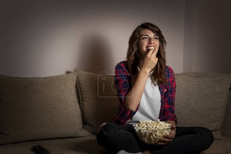 Téléchargez les photos : Belle jeune femme assise dans le noir sur un canapé du salon, s'amusant à regarder la télévision et à manger du pop-corn - en image libre de droit