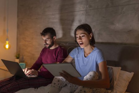 Téléchargez les photos : Beau jeune couple assis côte à côte dans le lit la nuit, lire et regarder des émissions de télévision sur ordinateur portable et tablettes - en image libre de droit