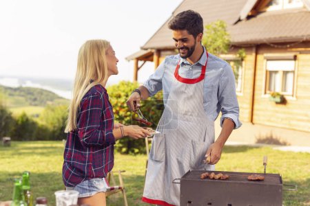 Téléchargez les photos : Couple faisant une fête barbecue arrière-cour par une journée d'été ensoleillée, griller de la viande et s'amuser - en image libre de droit