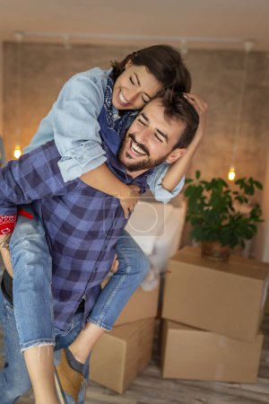 Téléchargez les photos : Jeune couple emménageant ensemble, s'amusant tout en déballant des boîtes en carton, copain piggyback sa petite amie et rire - en image libre de droit