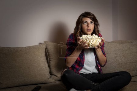 Téléchargez les photos : Belle jeune femme assise dans le noir sur un canapé du salon, effrayée, regardant un film d'horreur à la télévision et mangeant du pop-corn - en image libre de droit