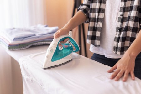 Photo for Detail of female hands holding the iron, ironing washed, wrinkled clothes on the ironing board - Royalty Free Image