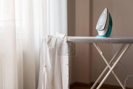 Photo for Ironed shirt hanging from an ironing board next to a hot iron. Selective focus on the iron - Royalty Free Image