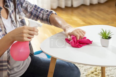 Foto de Hermosa mujer joven limpiando el polvo de una mesa de centro de la sala de estar con un spray limpiador y un paño; asistente de limpieza limpiando el polvo - Imagen libre de derechos