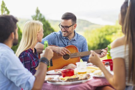 Foto de Grupo de amigos haciendo una fiesta de barbacoa al aire libre, tomando cerveza, asando carne, tocando la guitarra, cantando y divirtiéndose - Imagen libre de derechos