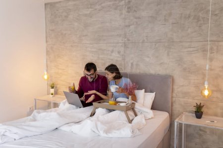 Foto de Hermosa pareja joven enamorada usando pijamas sentados en la cama por la mañana, desayunando, tomando café y leyendo noticias usando computadora portátil - Imagen libre de derechos