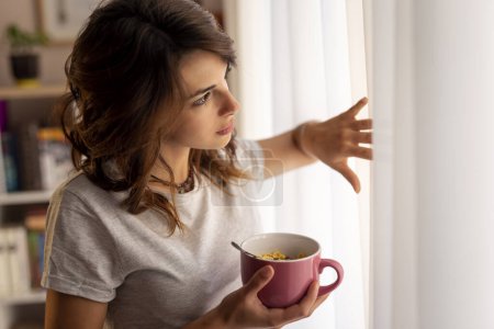 Téléchargez les photos : Belle jeune femme en pyjama, debout à côté de la fenêtre du salon, tenant un bol de céréales pour le petit déjeuner et profitant du week-end matin - en image libre de droit