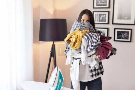 Bored and annoyed young woman holding bunch of washed and dried, wrinkled clothes ready for ironing; woman and home chores