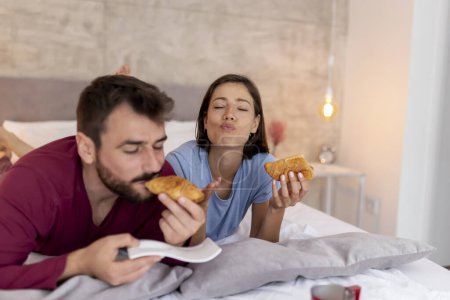 Foto de Hermosa pareja joven enamorada usando pijamas, acostada en la cama, desayunando después de despertarse por la mañana - Imagen libre de derechos
