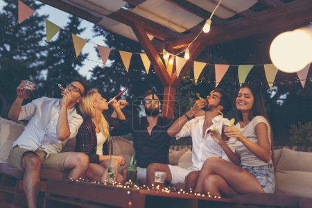 Foto de Grupo de jóvenes amigos soplando silbatos de fiesta, bebiendo cerveza y divirtiéndose en una fiesta de verano al aire libre - Imagen libre de derechos