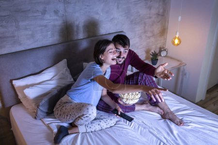 Photo for Beautiful young couple in love, sitting in bed side by side, hugging, eating popcorn and having fun watching a movie at night - Royalty Free Image
