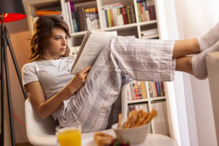 Foto de Hermosa joven con pijama, sentada junto a la ventana de la sala de estar, desayunando y leyendo los periódicos - Imagen libre de derechos