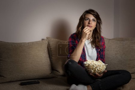 Foto de Hermosa joven sentada en la oscuridad en un sofá de la sala de estar, divirtiéndose viendo la televisión y comiendo palomitas de maíz - Imagen libre de derechos