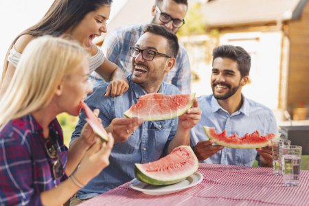Téléchargez les photos : Groupe d'amis déjeuner en plein air, manger des tranches de pastèque fraîche et s'amuser - en image libre de droit