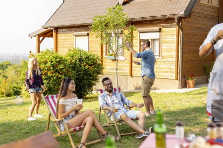 Téléchargez les photos : Groupe d'amis faire une fête barbecue dans la cour, griller de la viande, boire de la bière, jouer au badminton et s'amuser par une journée ensoleillée d'été - en image libre de droit