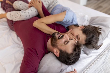 Téléchargez les photos : Couple amoureux couché au lit après le réveil, étreignant, riant et appréciant leur temps ensemble, femme chuchotant un secret à l'oreille de l'homme - en image libre de droit