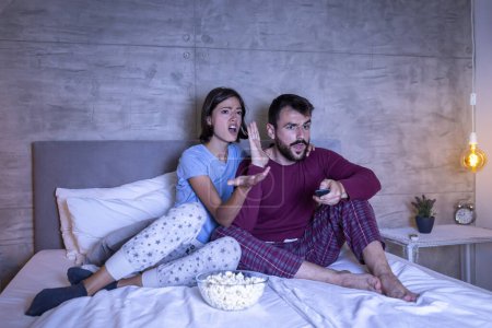 Téléchargez les photos : Couple portant un pyjama assis dans le lit, se battant à propos de la télécommande et le choix des programmes de télévision à regarder, homme changer de chaîne tandis que la femme est en colère, criant - en image libre de droit