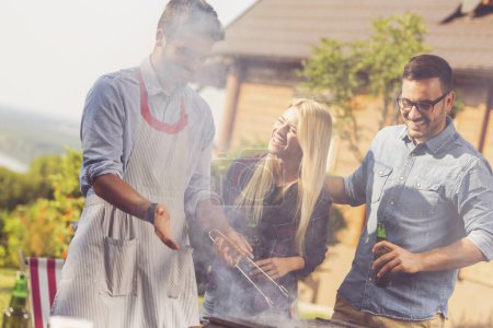 Foto de Grupo de amigos haciendo una fiesta de barbacoa en el patio trasero, asando carne, bebiendo cerveza y divirtiéndose en un día soleado de verano - Imagen libre de derechos