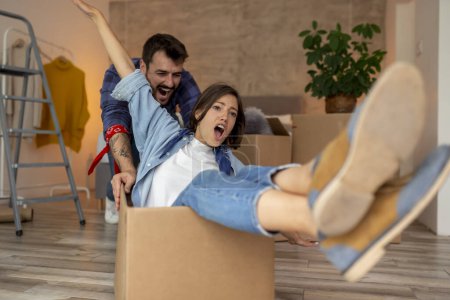 Photo for Young couple in love moving in together, having fun while unpacking cardboard boxes with their belongings, guy pushing girl around in a cardboard box - Royalty Free Image