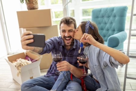 Photo for Beautiful young couple in love moving in new apartment, drinking wine and having fun taking selfies with the keys of their new home - Royalty Free Image
