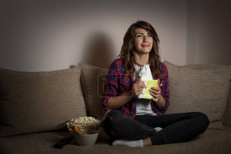Téléchargez les photos : Belle jeune femme assise dans le noir sur un canapé du salon, regardant un film dramatique et sanglotant - en image libre de droit
