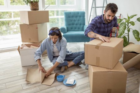 Photo for Couple in love packing their possessions into cardboard boxes, getting ready for relocation - man taping boxes using packing machine while woman is wrapping fragile things into paper - Royalty Free Image