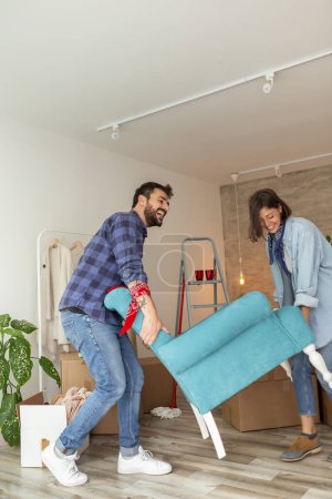 Photo for Couple in love moving in together, having fun while carrying an armchair and setting up the new furniture arrangement - Royalty Free Image