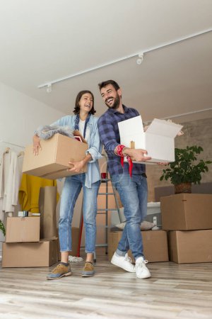 Photo for Couple in love moving in together, having fun while carrying cardboard boxes filled with their possessions and setting up the new apartment - Royalty Free Image