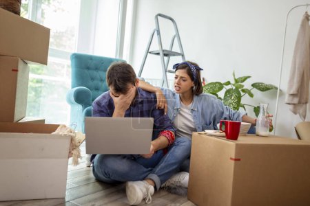 Photo for Beautiful young couple in love moving in together, sitting among cardboard boxes in their new apartment, worried and annoyed, having trouble with bank loan - Royalty Free Image