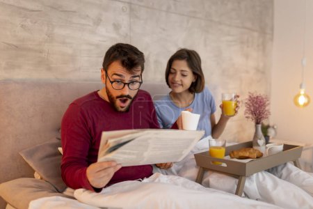 Foto de Pareja joven enamorada vistiendo pijamas, sentada en la cama por la mañana, leyendo periódicos y desayunando, relajándose en casa - Imagen libre de derechos