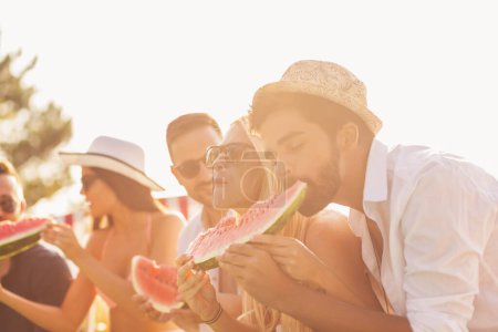 Téléchargez les photos : Groupe d'amis à une fête d'été au bord de la piscine, assis au bord d'une piscine, mangeant des tranches de pastèque froide et s'amusant - en image libre de droit