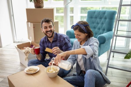 Foto de Hermosa pareja joven enamorada de mudarse juntos, sentados entre cajas de cartón en su nuevo apartamento, divirtiéndose mientras desayunan - Imagen libre de derechos