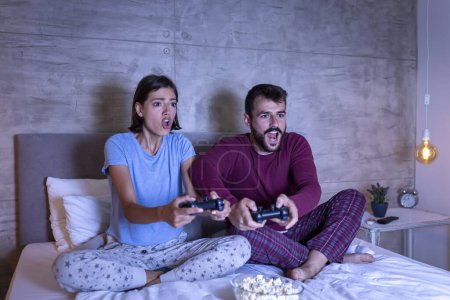 Téléchargez les photos : Beau jeune couple en pyjama, s'amuser à la maison, jouer à des jeux vidéo au lit la nuit, femme excitée après avoir gagné le match, homme déçu après avoir perdu - en image libre de droit