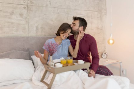 Foto de Hermosa joven pareja enamorada usando pijamas sentada en la cama, hombre sorprendente mujer trayendo su desayuno y flores en la cama en una bandeja, besándola en la frente - Imagen libre de derechos