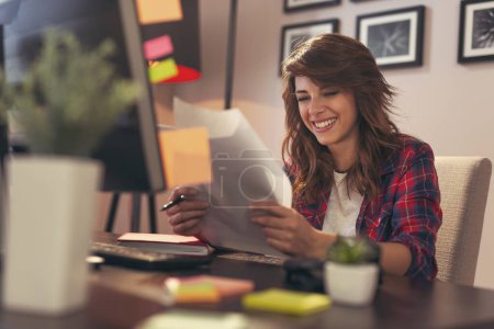 Foto de Mujer joven sentada en un escritorio en su oficina en casa, sosteniendo documentos alegres después de un éxito empresarial - Imagen libre de derechos