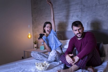 Foto de Hermosa pareja joven usando pijamas, divirtiéndose en casa, jugando videojuegos en la cama por la noche, mujer emocionada después de ganar el juego, hombre decepcionado después de perder - Imagen libre de derechos